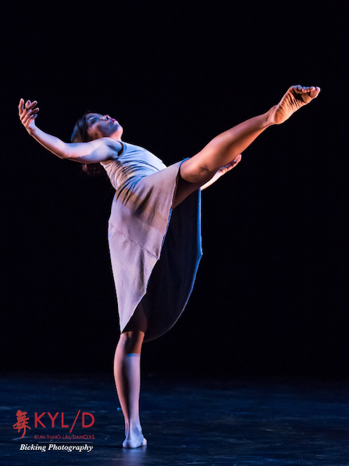 A female dancer in a long dress. Balances on one leg as her right leg is extended high in front of her, her torso arches back and her head looks to the sky